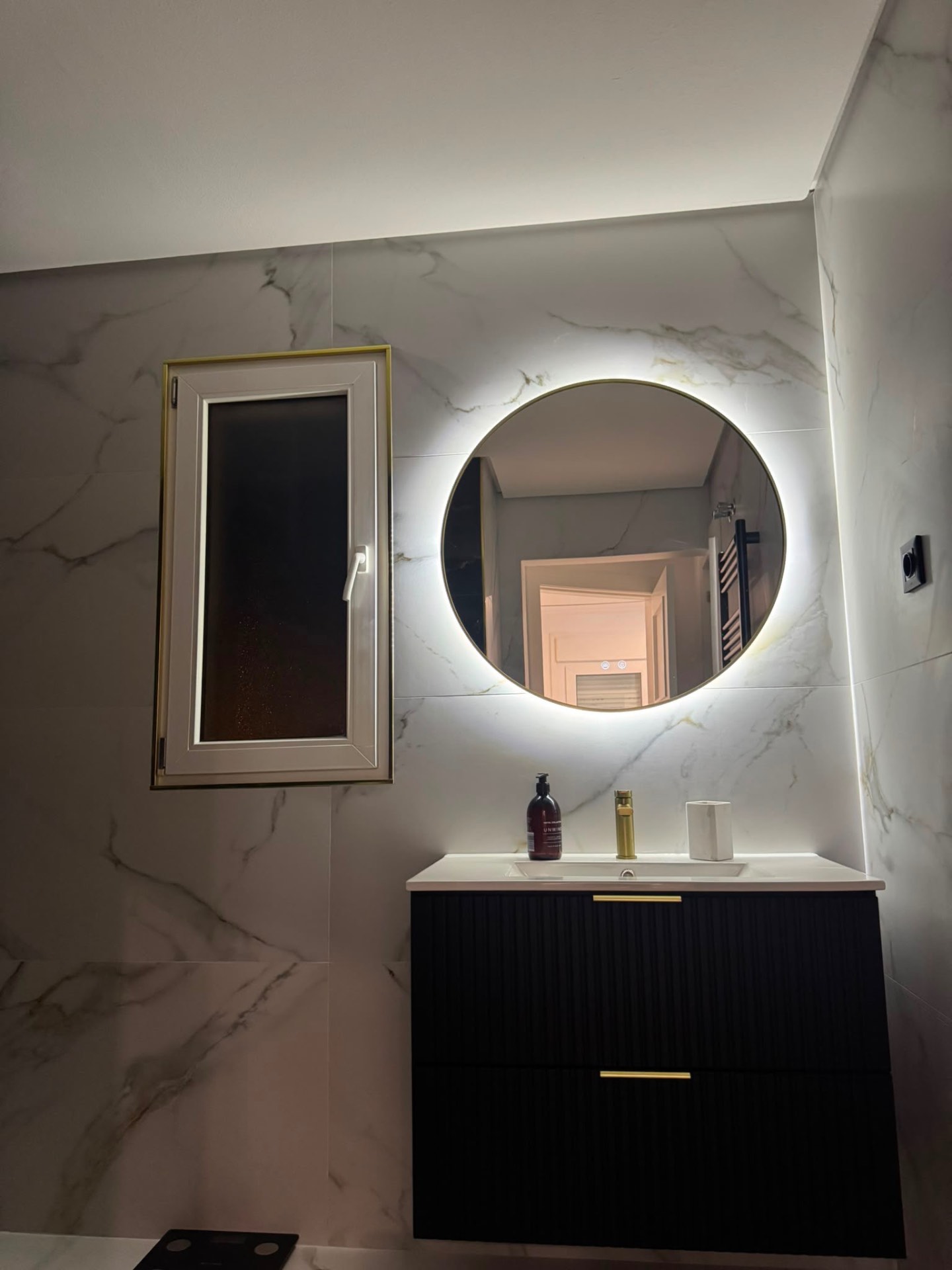Fluted black vanity unit with brass handles under a round backlit LED mirror on marble wall